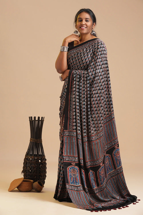 Full-length studio portrait of a model gently posing while showcasing the classic black modal silk saree with rich contrasting borders and fluid drape