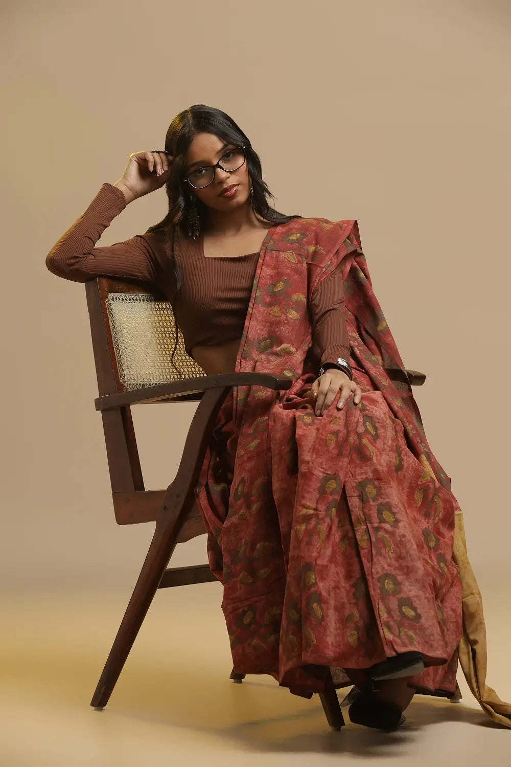 Crimson Earth red, brown and yellow hand-block Bagru cotton saree displayed by a model seated beside a wooden chair