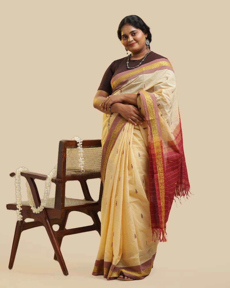 Full-length studio view of a model wearing the ivory cream Kasaragod cotton saree with a contrasting maroon pallu, posed next to a wooden chair with a decorative garland