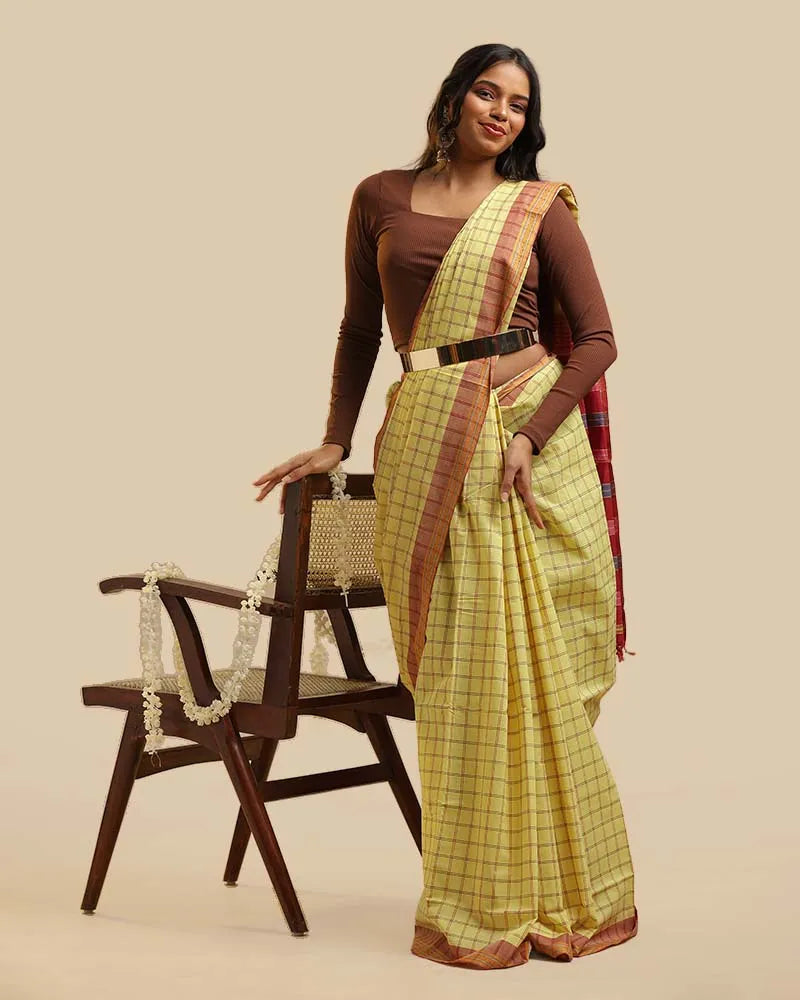 Full-length studio view of a model wearing the meadow yellow Kasaragod cotton saree with a checked body and rich maroon pallu, posed next to a wooden chair with a white garland
