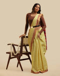 Full-length studio view of a model wearing the meadow yellow Kasaragod cotton saree with a checked body and rich maroon pallu, posed next to a wooden chair with a white garland