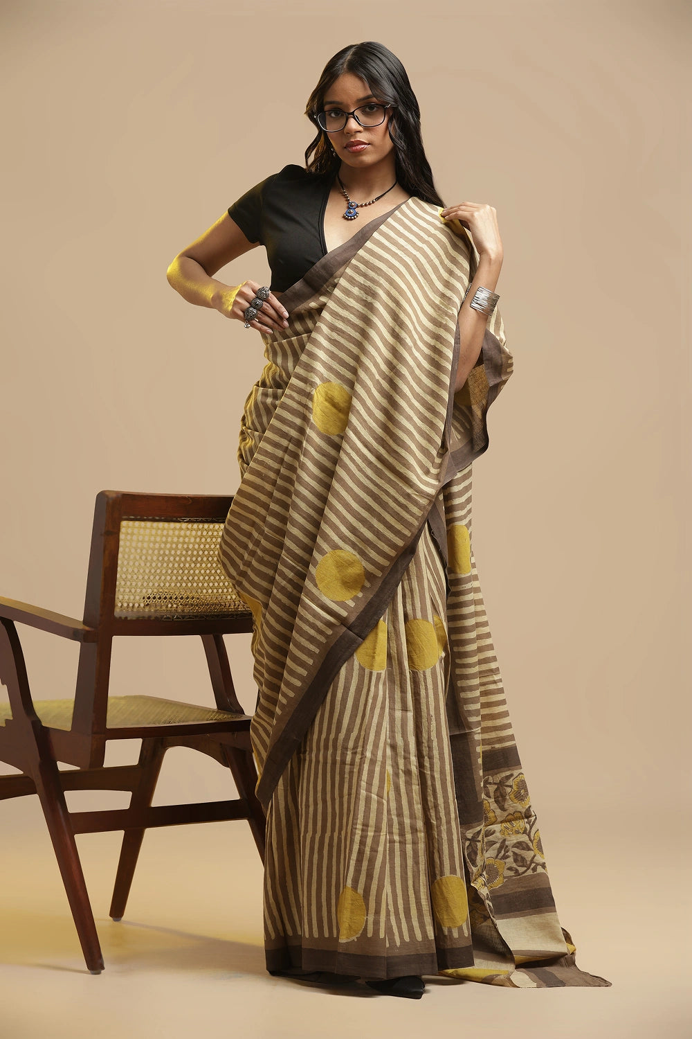 Modern Minimalism beige, brown and yellow Bagru cotton saree featuring bold stripes and polka dots displayed by a model posing beside a wooden chair