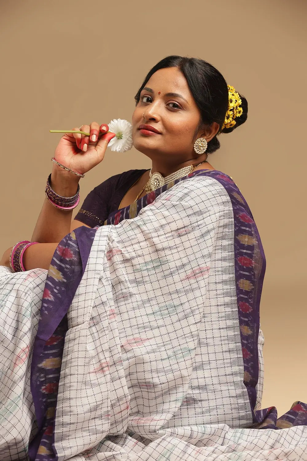 White and purple Muthyampet check cotton saree featuring pearl ikat patterns displayed by a seated model holding a white flower