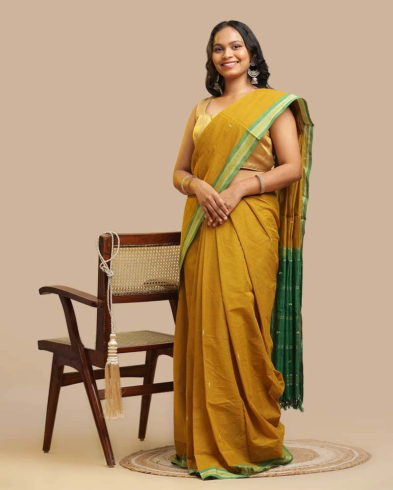 Olive Green Udupi Cotton Saree with emerald green striped border and woven buttas worn by a model standing next to a wooden chair