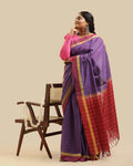 Full-length studio view of a model wearing the royal purple Kasaragod cotton saree with a deep maroon pallu, posed next to a wooden chair with a white garland