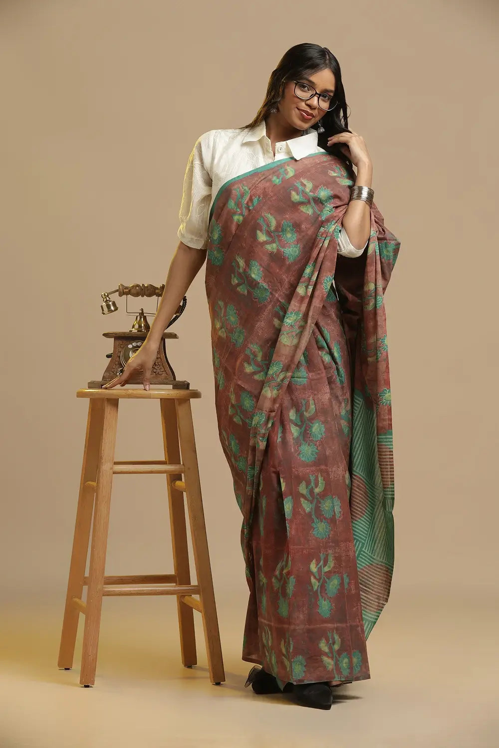 Shades of Earth brown and green handblock print cotton saree displayed by a model posing beside a wooden stool