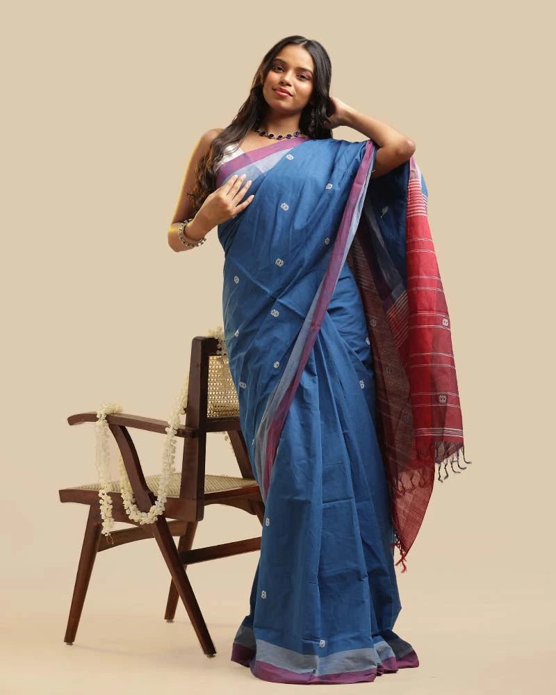 Full-length studio view of a model wearing the twilight blue Kasaragod cotton saree with a contrasting maroon pallu, posed next to a wooden chair with a white garland